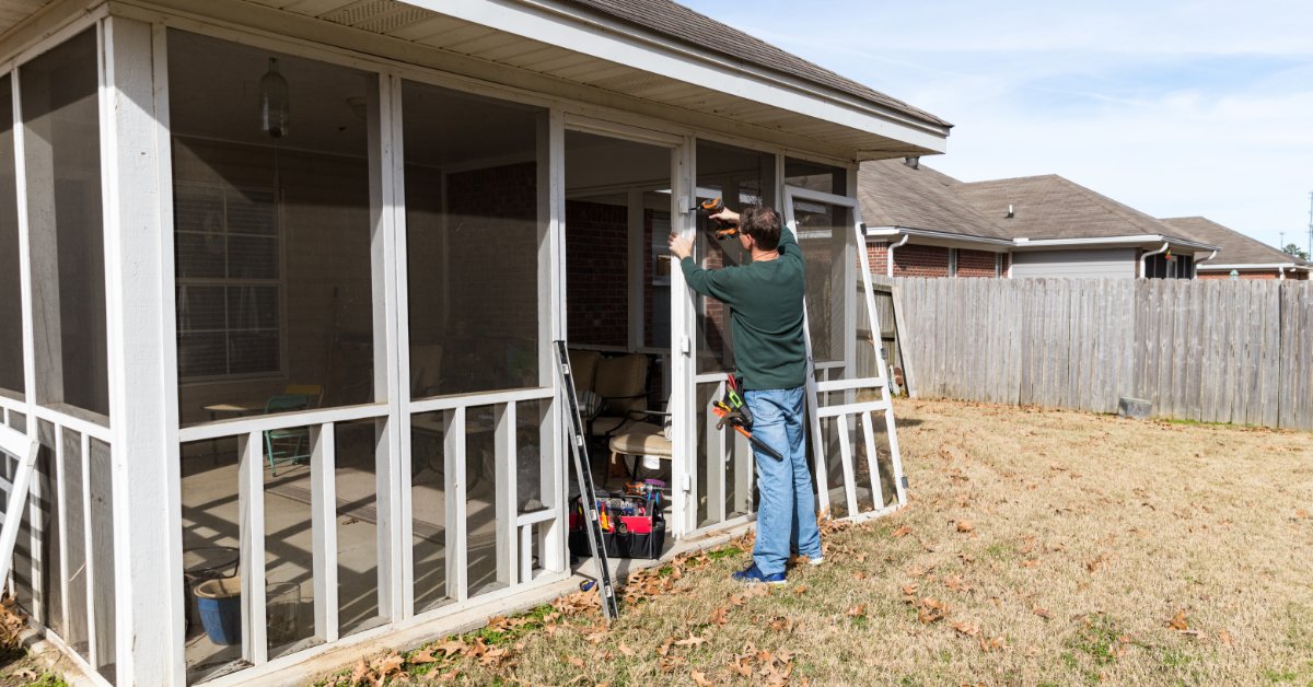 Screened In Porch Upgrades That Add Home Value A man repairing a screened porch door beside ladders, hand tools, white posts, dry grass, and a wood fence.