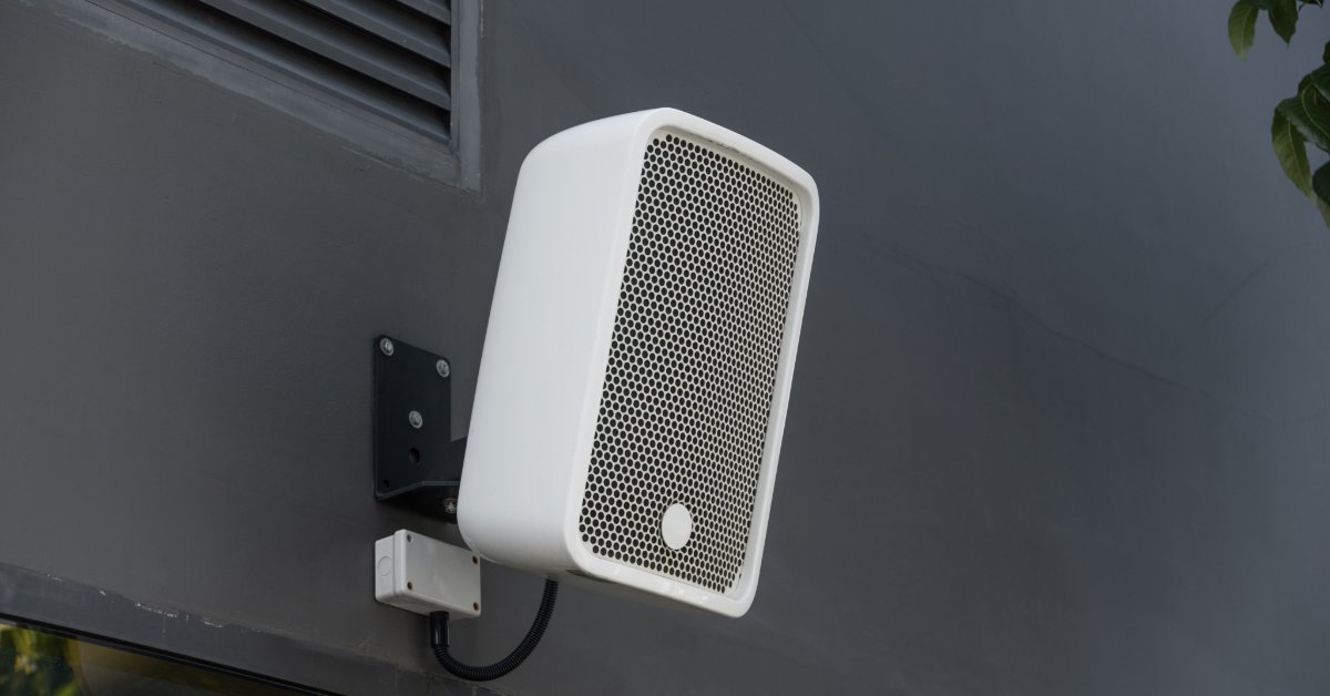 Screened In Porch Upgrades That Add Home Value A white outdoor speaker mounted on a dark wall near a vent, cables, window glass, and nearby green leaves.