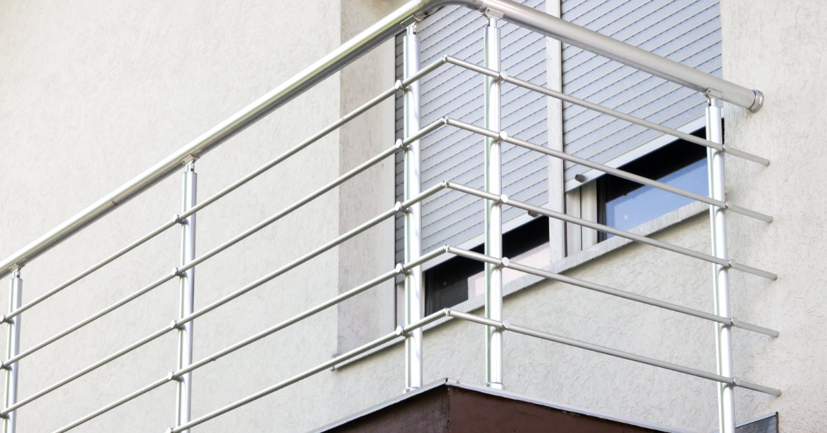 Aluminum Railings That Blend Strength and Style A silver metal balcony railing with horizontal bars attached to a beige building beside a window with closed shutters.