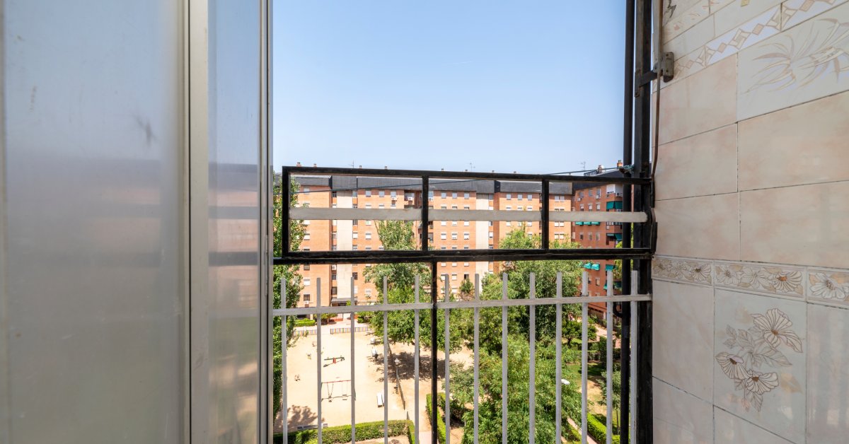 Aluminum Railings That Blend Strength and Style A small balcony with a metal railing, a tall cabinet, tiled walls, apartment buildings, trees, and courtyard paths.