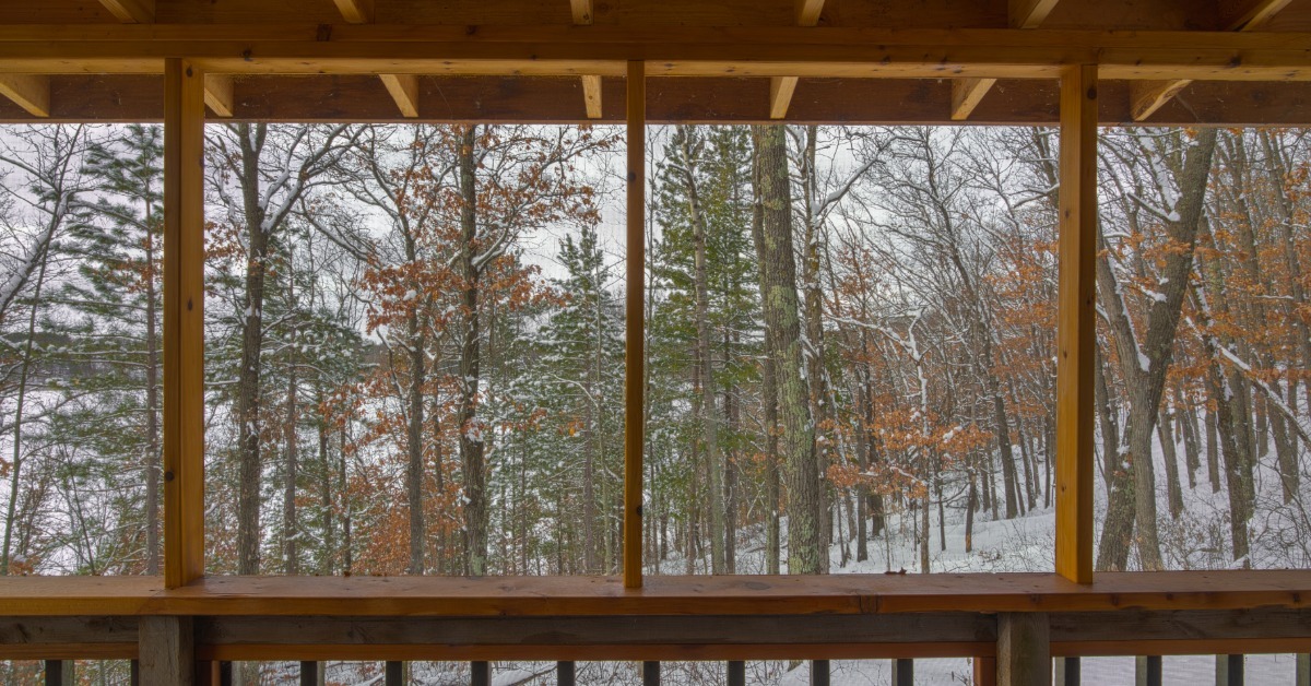 A wooden screened porch with thick beams, vertical posts, and a lower railing overlooking snowy woods.