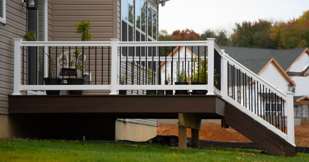 Expert Guide to Choosing Quality Railing Parts A raised deck with white railing, stairs, potted plants, grass, and beige siding on a house exterior.