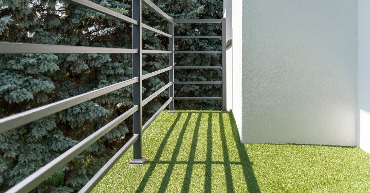 A narrow balcony with metal railings, green synthetic grass flooring, tree branches, and bright sunlight.
