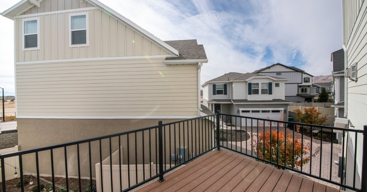 A wooden house deck with black metal railings, nearby homes, shrubs, and a partly cloudy sky above the yard.