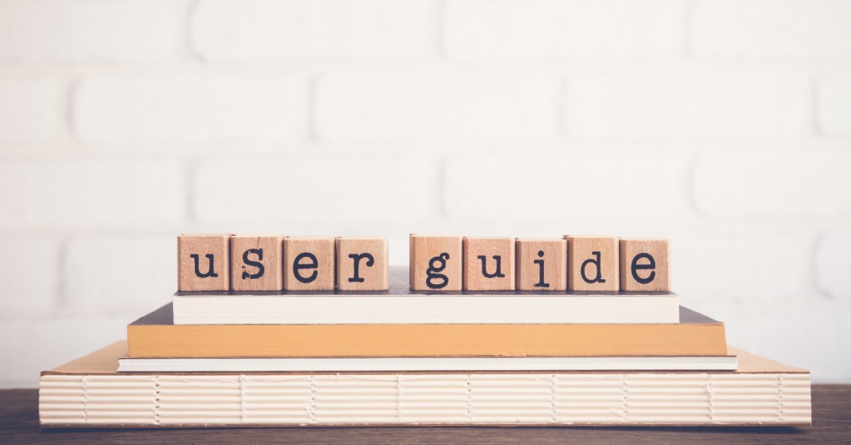 Wooden letter blocks spelling user guide resting on stacked books before a pale brick wall background.