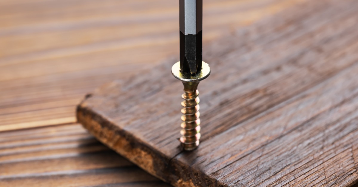 A screwdriver bit pressing a gold screw into a dark wooden board lying on a lighter wooden work surface.