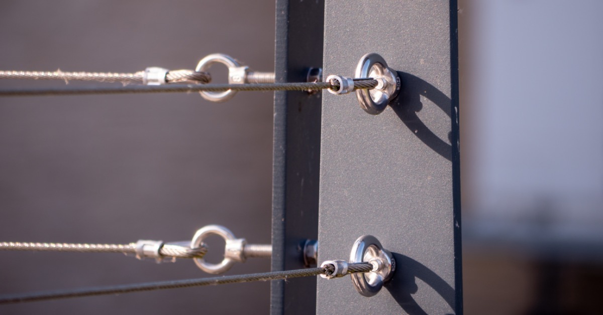 Steel cables and circular metal fittings extending horizontally through dark rectangular railing posts in sunlight.