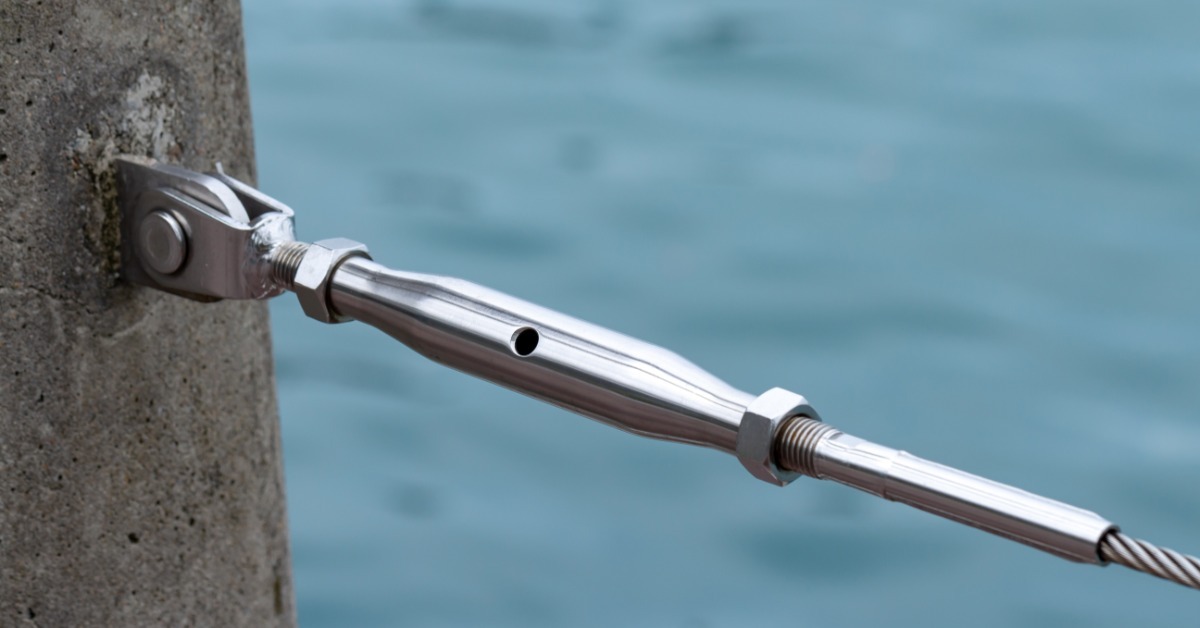 A stainless steel cable tensioner with threaded fittings connecting a steel cable to a concrete post beside blue water.
