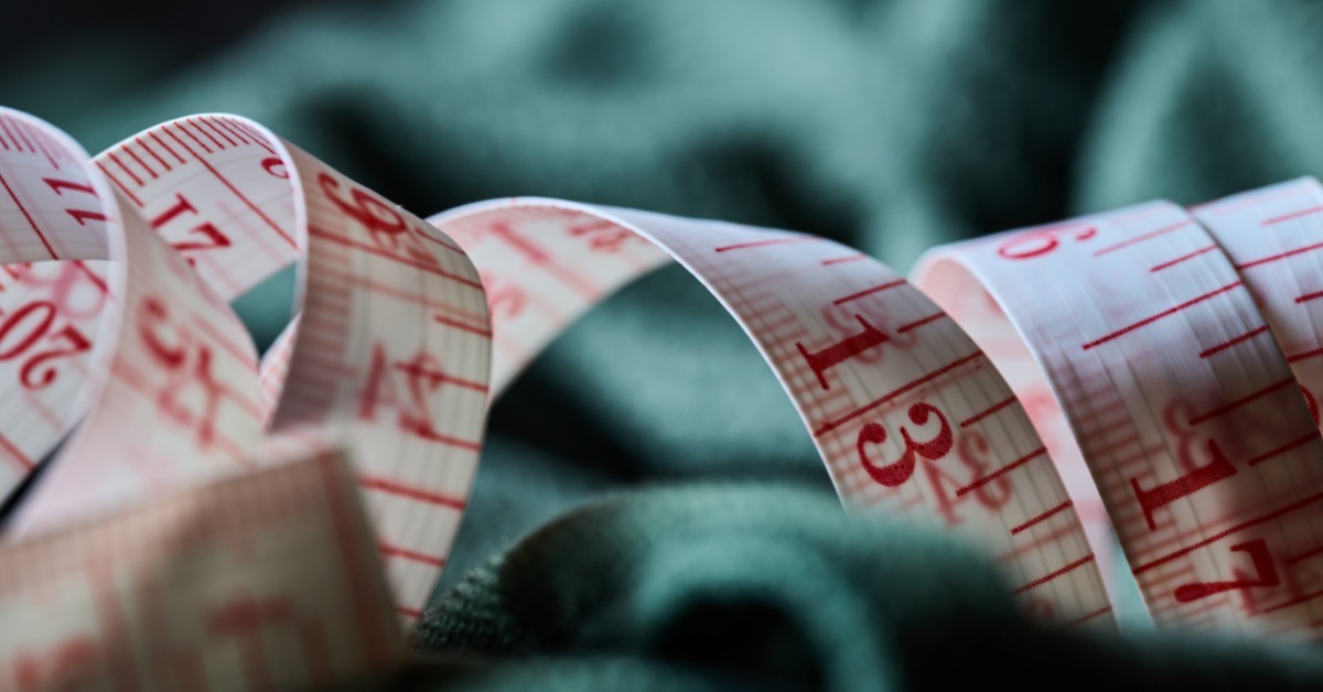 A white measuring tape with red markings curling across textured green fabric in overlapping loops and folds.