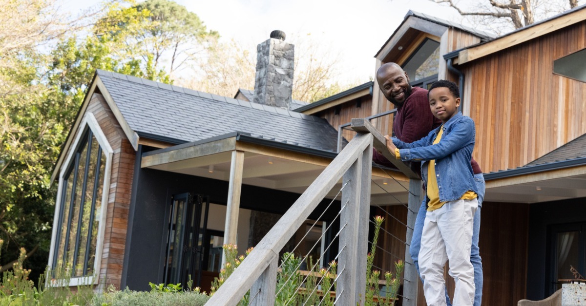 A man and boy standing together on wooden porch stairs and leaning against a cable railing beside a house.