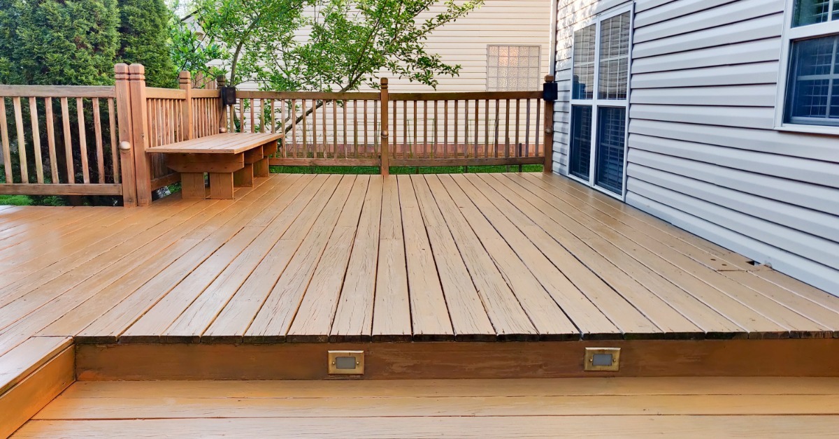 A large wooden deck with broad steps, built-in lights, a railing, and a bench beside a house with gray siding.