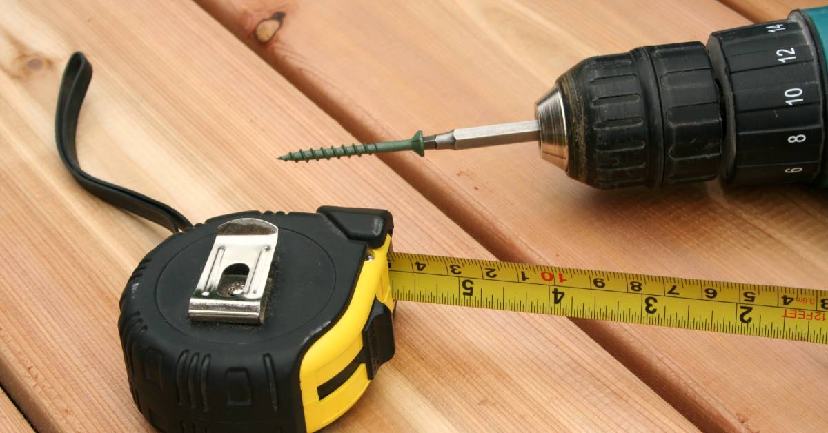 A drill holding a screw above wood boards, with a black and yellow tape measure stretched across the surface.