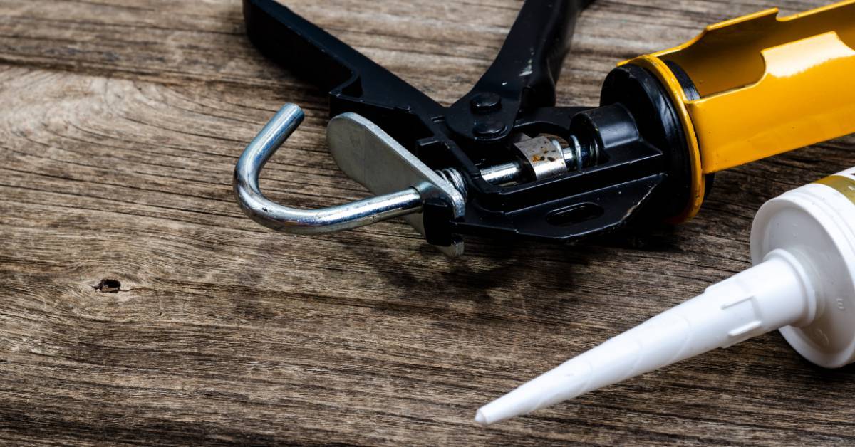 A yellow and black caulking gun with a white tube lying on weathered wooden planks near a metal hook and handle.