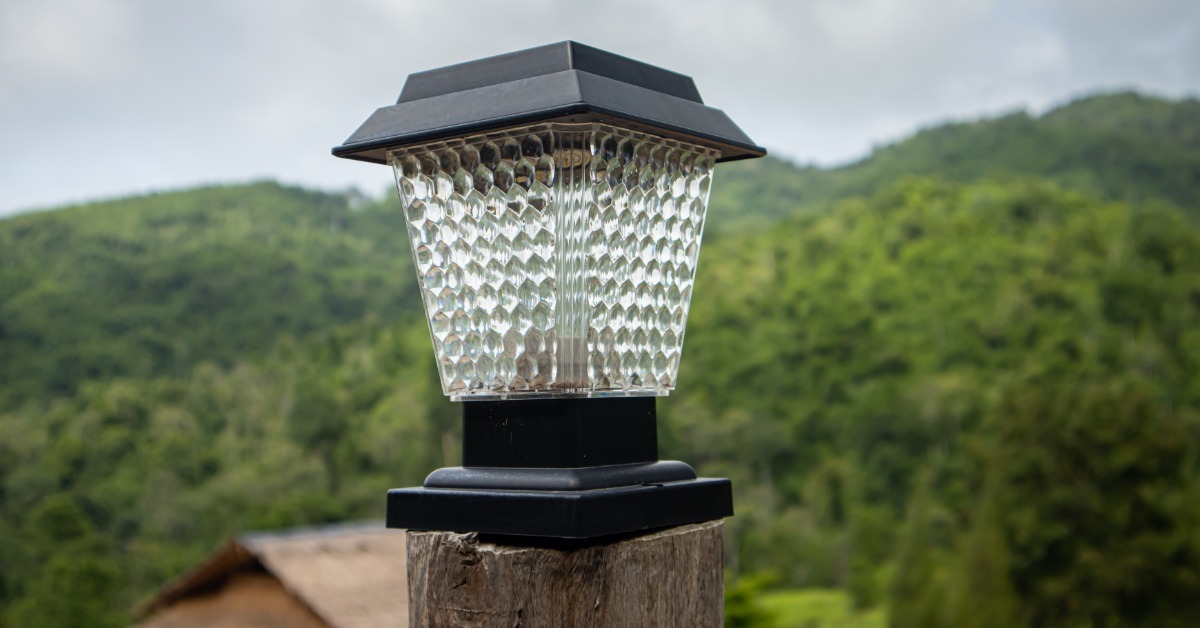 A black solar lamp on a wooden post standing before green hills, trees, a cloudy sky, and a small roof.