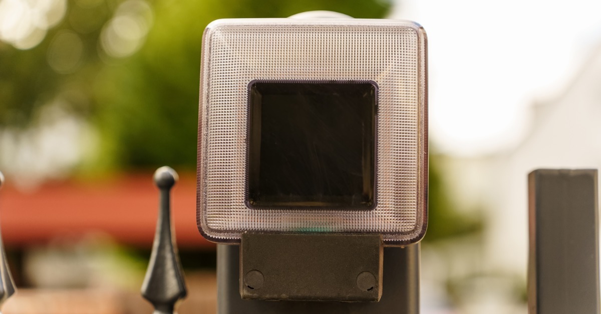 A square solar light attached to a black fence post with trees, a red roof, and buildings in the background.