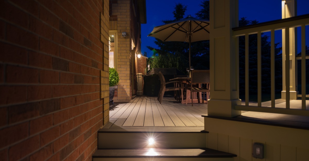 A lit deck walkway running beside a brick wall at night, leading toward outdoor dining under a patio umbrella.