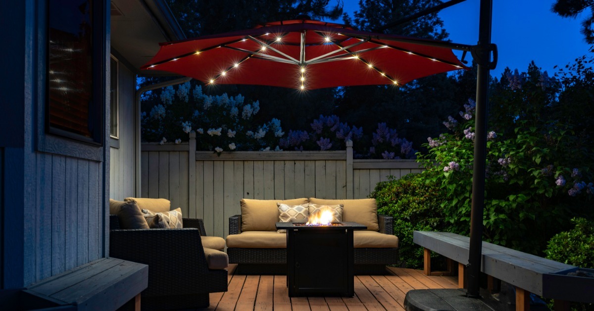 A backyard deck at twilight showing outdoor seating gathered near a glowing fire table beneath a lit umbrella.