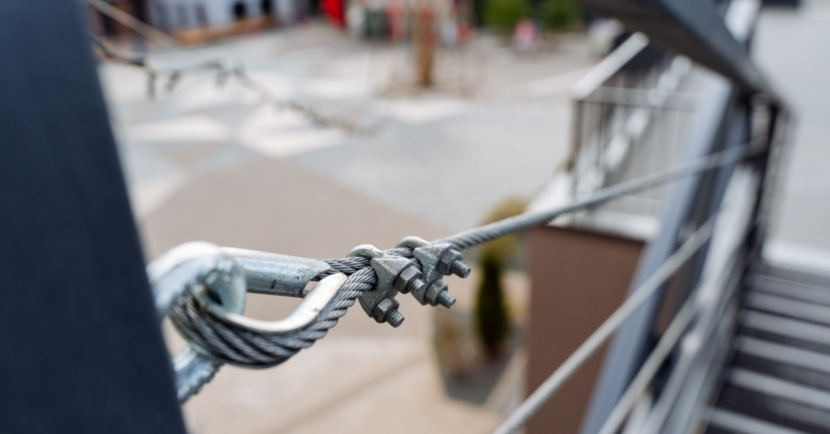 A braided steel cable secured to a metal railing post with a looped fitting and bolted clamps beside outdoor stairs.