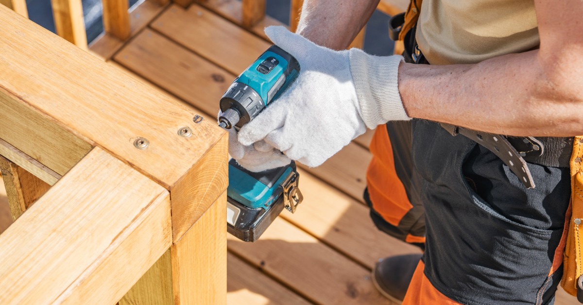 A man using a power drill to put together two parts of a wooden railing on his deck. He is wearing white gloves.