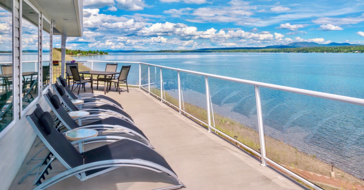 A deck with a glass railing and multiple beach chairs. The deck overlooks a large lake. There is also a table and chairs nearby.