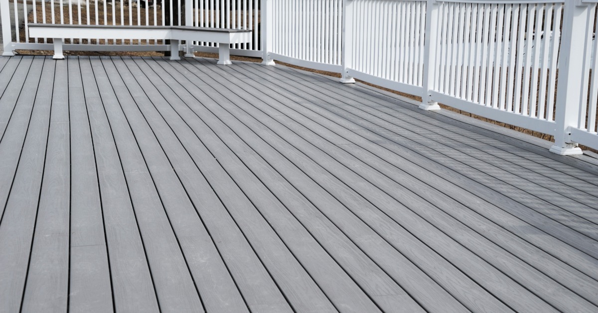 A gray deck with a white railing surrounding it. There is also a gray and white bench sitting at the end of the deck.