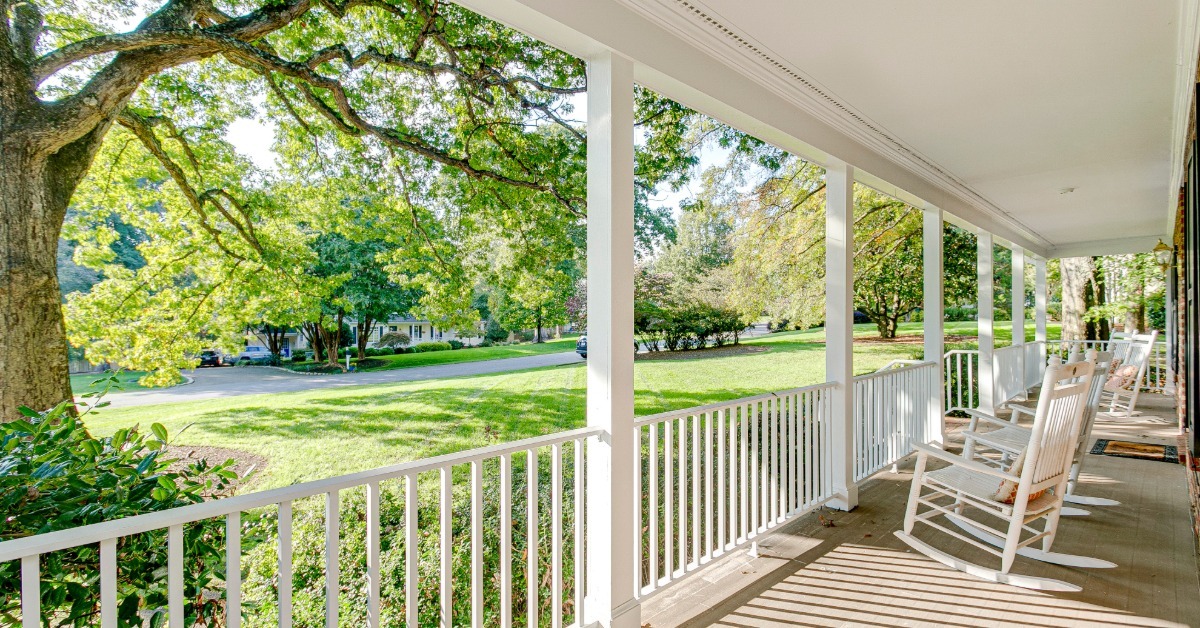 The view of the street from a home's front porch. There are a few white rocking chairs sitting out front.