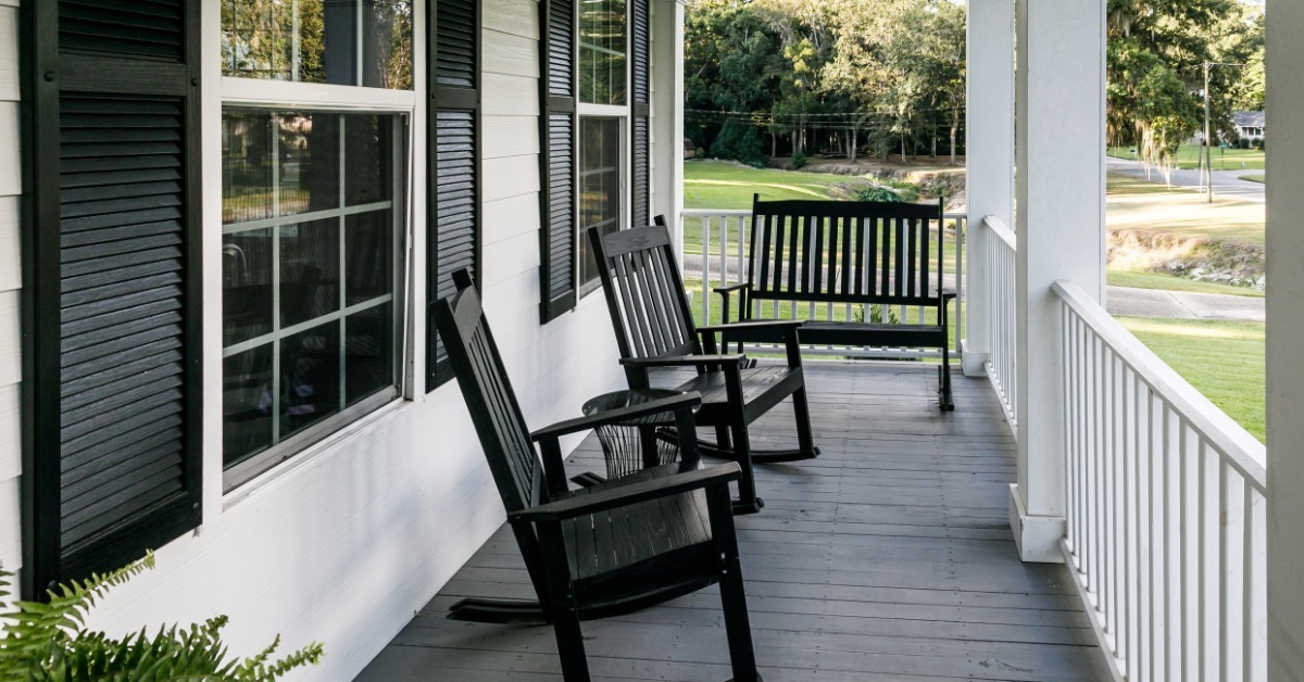 Two black rocking chairs and a bench all sitting on a home's front porch. There is a small table between the chairs.