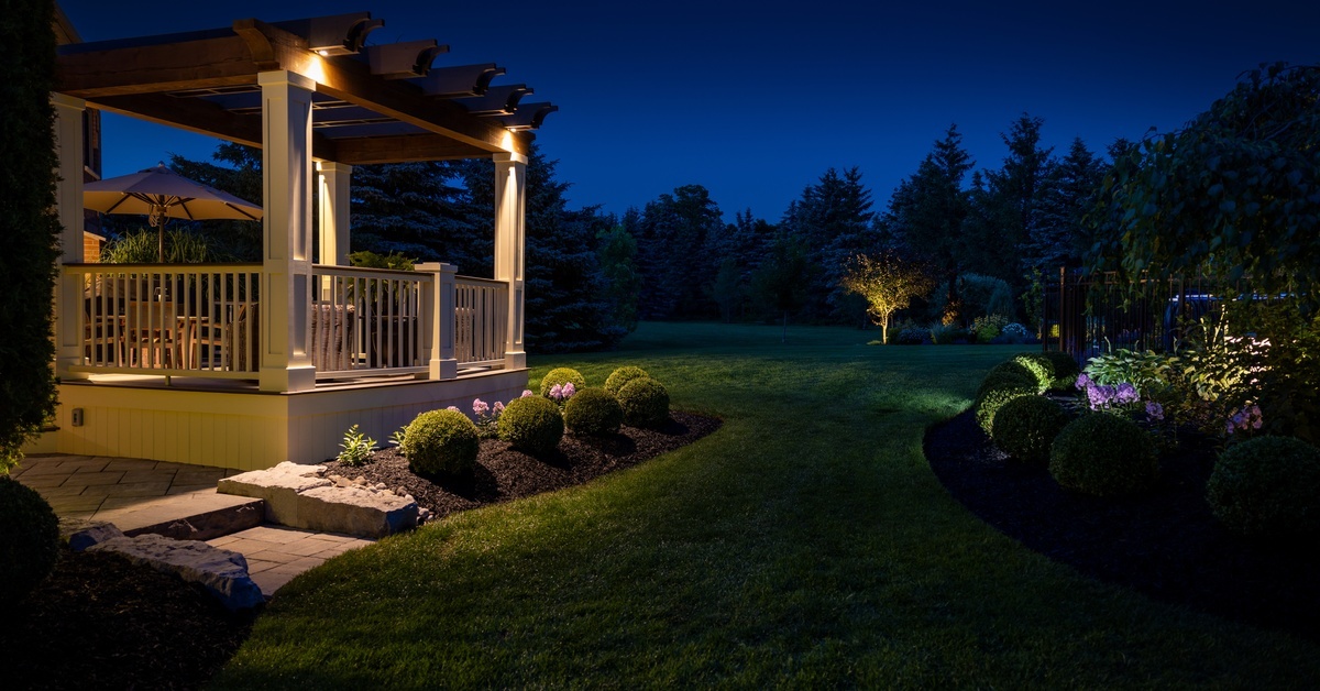 A lit-up deck next to a well-maintained yard. There are lights in the yard illuminating the trees and bushes.
