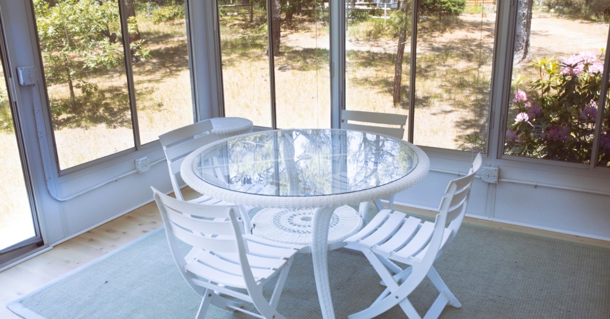A glass table surrounded by four white chairs. This dining setup is sitting inside a screened-in porch.