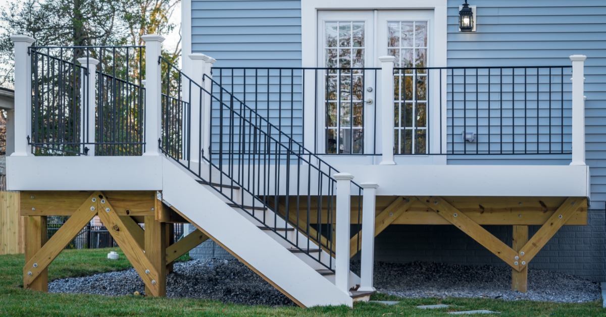 A classic backyard deck with black metal railings and white columns connected to a small home with blue siding.