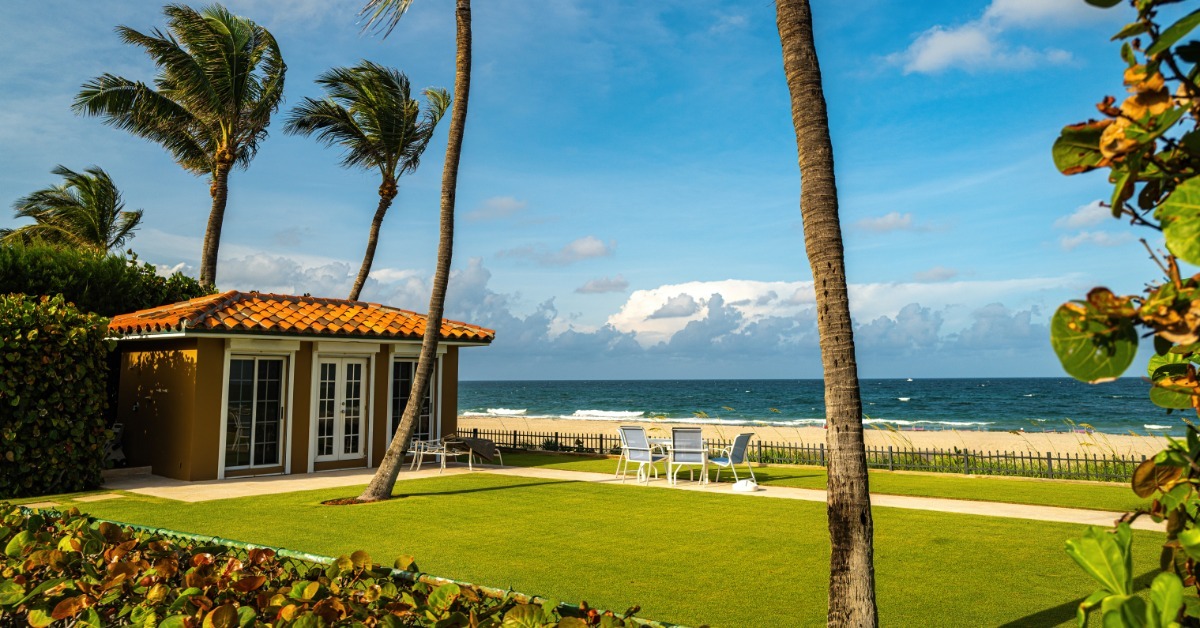 A tropical beach house with ocean views and palm trees. There is a table and some chairs facing the beach.