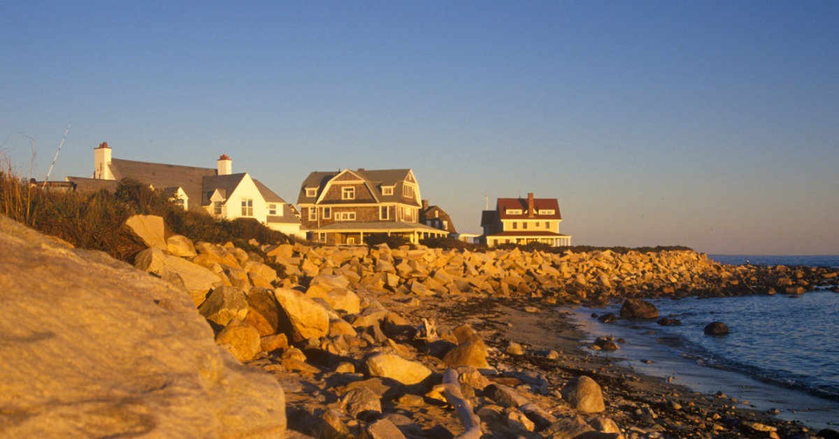 A few oceanfront homes along a rocky shoreline at sunset, with waves coming in during the warm evening light.
