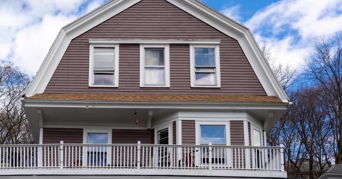 A single-family home with a gambrel roof and balcony. There is a cloudy, blue sky behind the house.