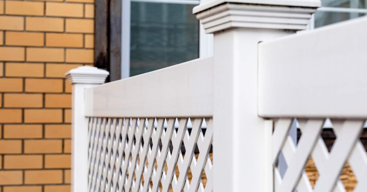 A white vinyl railing section with posts in front of a brick house. There is a window behind the railing.