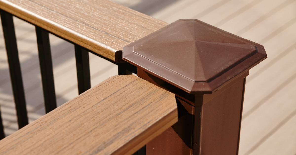 A close-up of a wooden deck railing with black balusters and a brown post. The sun is shining across the railing.