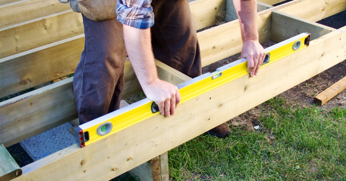 A person checks a wooden deck frame with a yellow spirit level while standing inside of the deck frame.