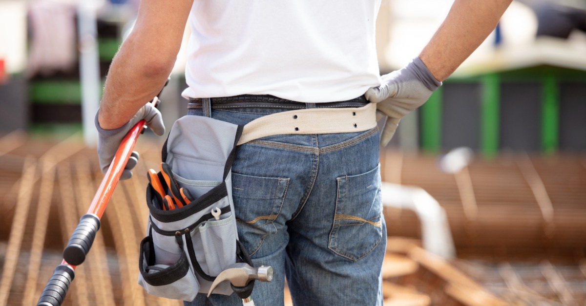 A person in jeans and work gloves wearing a tool belt while holding a long-handled tool at an outdoor worksite.