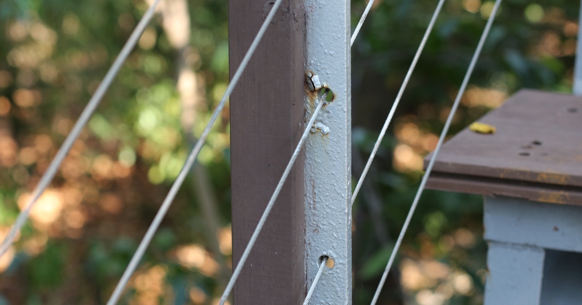 A metal railing post with peeling paint holding tensioned cables above a set of stairs for a wooden deck.