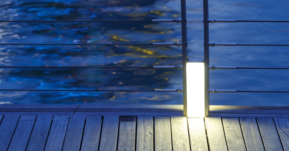 A stainless steel wire cable balustrade running along a deck that's beside a body of water at twilight.