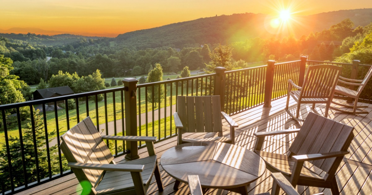  A wooden balcony facing wooded hills as sunlight sets over the hills. There is a table and chairs on the deck.