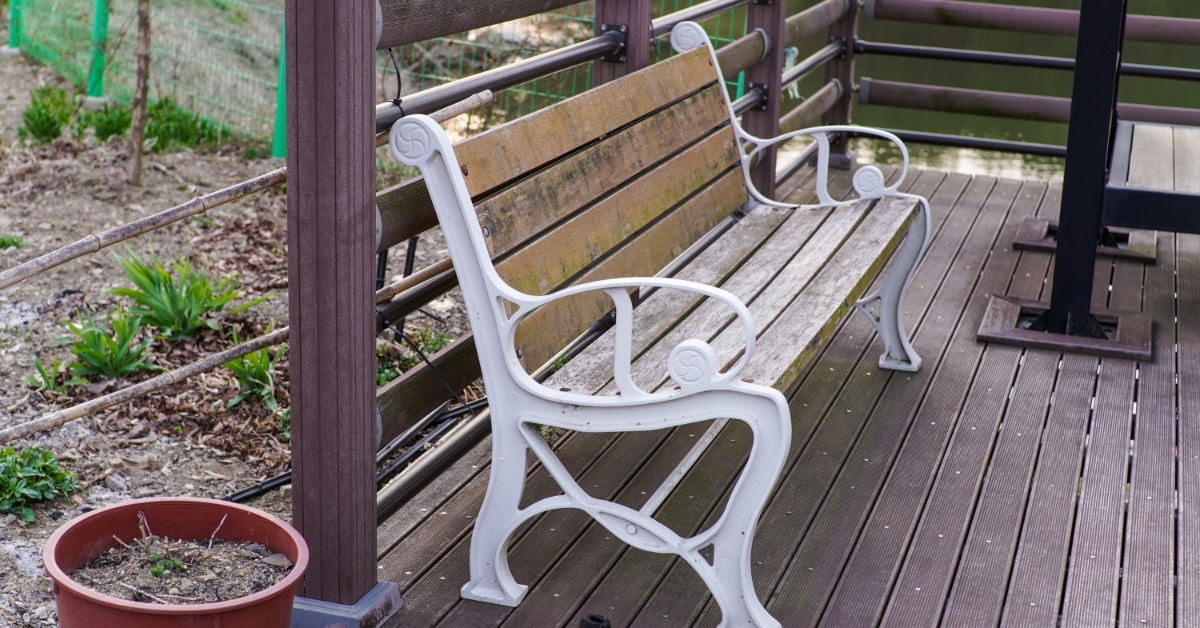 A weathered wooden bench sitting on a deck beside railing posts, with a pot full of dirt sitting next to the deck.
