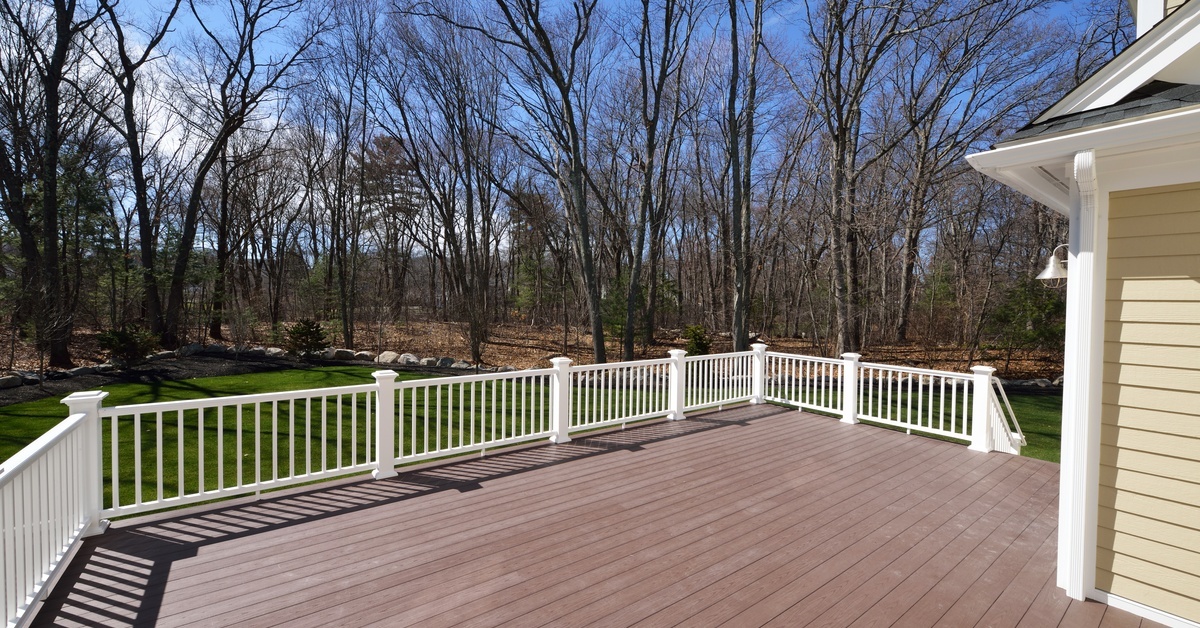 A large composite deck with white railings extending from a beige house toward a grassy yard and bare trees. A large composite deck with white railings extending from a beige house toward a grassy yard and bare trees.