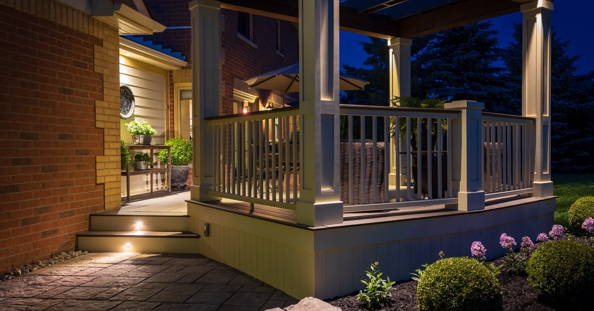 A well-lit deck and pergola beside a brick house, with railings, steps, shrubs, and outdoor seating at night.