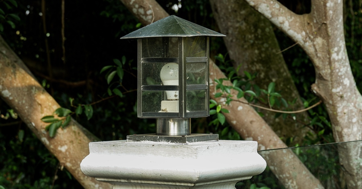 A weathered lantern sitting on a white post, with tree branches and green leaves not too far behind it.
