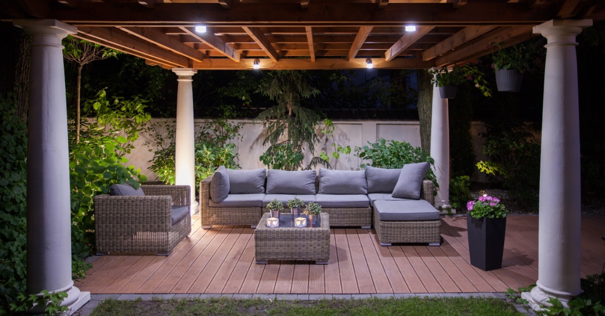 A covered patio outside a house at night, with wicker seating, columns, soft lighting, and a surrounding garden.
