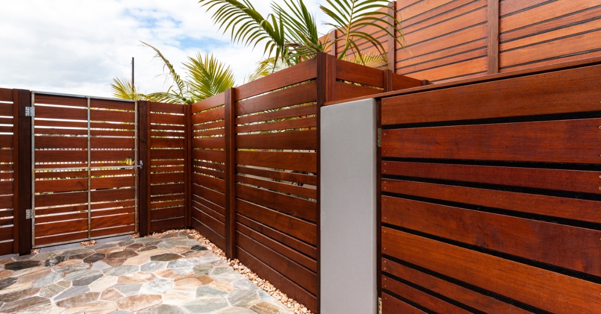 A modern backyard with horizontal wooden fencing, a side gate, stone flooring, and palm fronds above the walls.
