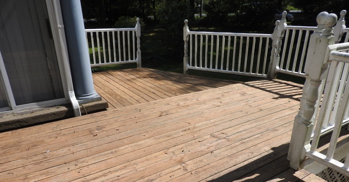A wooden deck with white railings beside a gray house, showing sunlit boards, shadows, and trees beyond.
